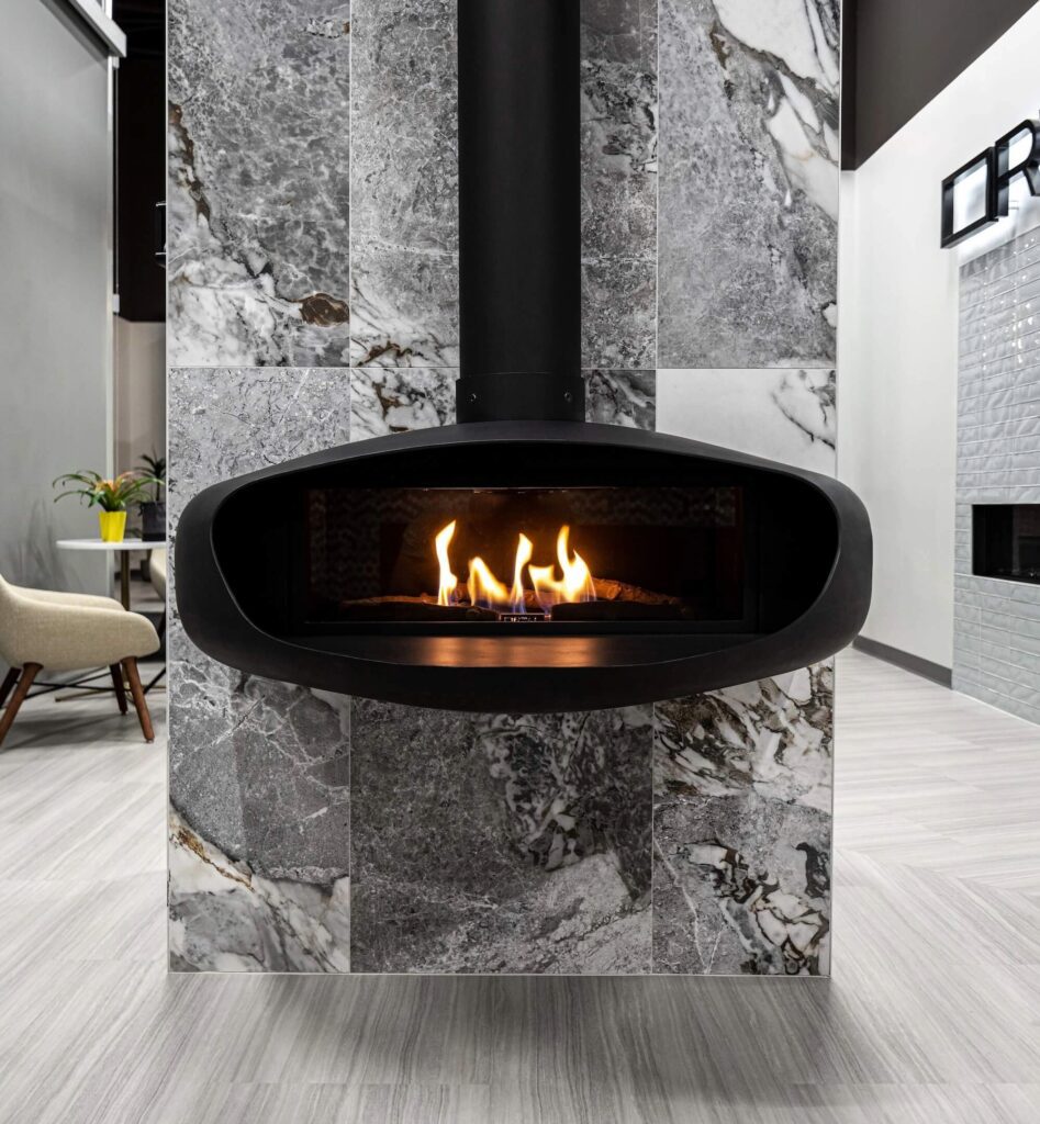 Modern suspended black fireplace with visible flames, set against a gray and white marble tile wall in a contemporary showroom.