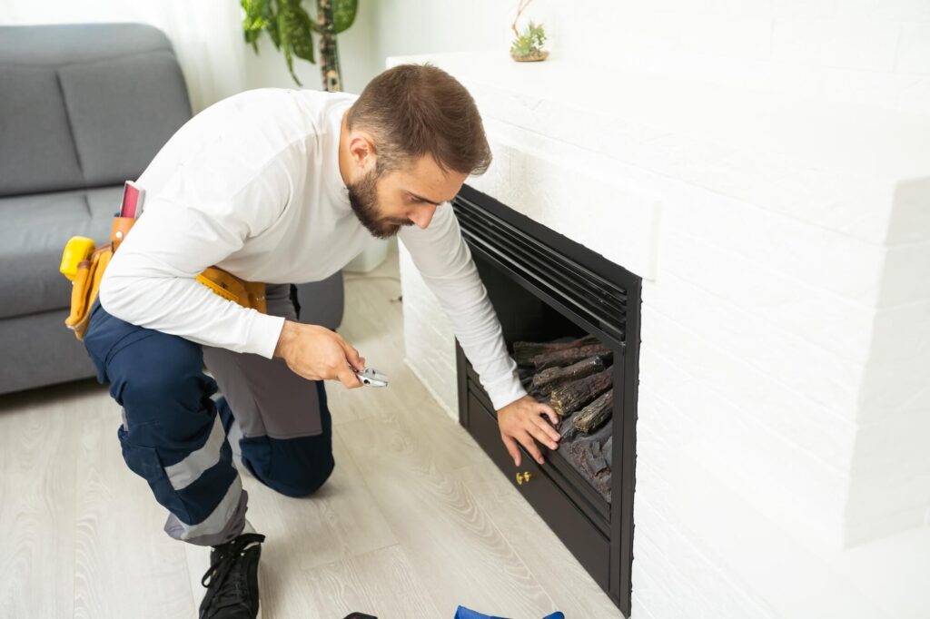 Technician kneeling in front of a fireplace, inspecting and adjusting gas logs with a tool belt at his side.